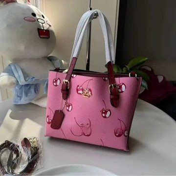 Pink tote bag printed with cherries with burgundy straps, beside a plush toy