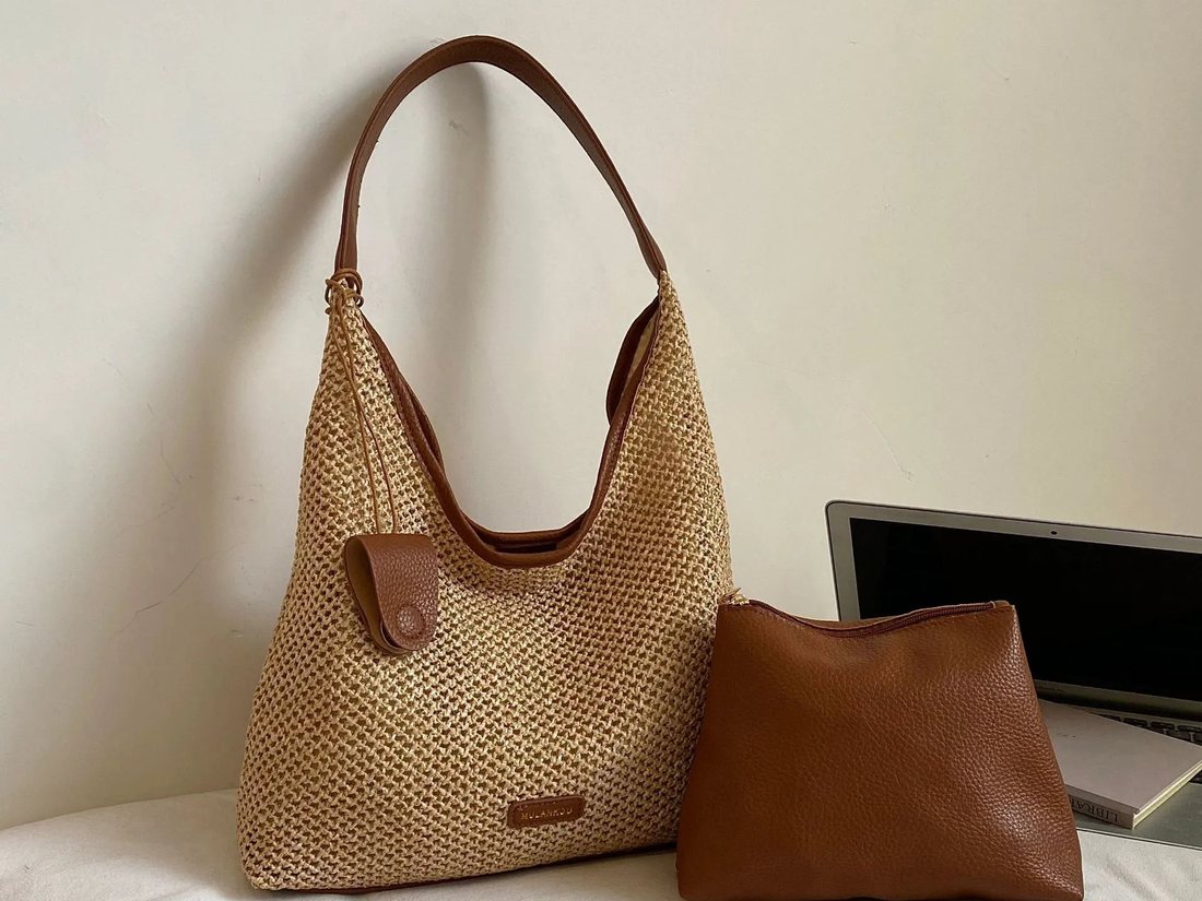 Woven straw shoulder bag with a brown strap standing next to a separate brown pouch on a bed, with a laptop behind